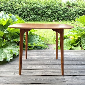 Puede incluir: Una mesa auxiliar rectangular de madera con un acabado liso de color marrón claro, que cuenta con cuatro patas cónicas y una superficie superior plana. La mesa presenta ensambladuras visibles donde las patas se unen al marco.
