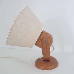 Może przedstawiać: Drewniana lampa biurkowa z beżowym materiałowym abażurem. Lampa ma okrągłą podstawę i obrotową głowicę, połączone drewnianymi wspornikami. Biały przewód zasilający wychodzi z podstawy.