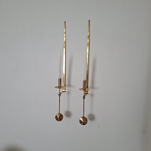 Puede incluir: Dos apliques de pared de latón con velas blancas. Cada aplique tiene un brazo largo y delgado con una base circular y un pequeño portavelas redondo. Los apliques están montados en una pared blanca, creando una estética minimalista y elegante.