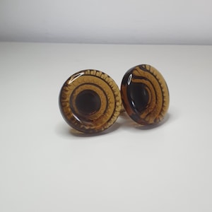 Könnte beinhalten: Zwei runde Ohrstöpsel aus bernsteinfarbenem Glas mit einem Spiraldesign. Die Stöpsel bestehen aus einem durchscheinenden Material, und das Design ist durch das Glas sichtbar. Sie werden auf einer weißen Oberfläche präsentiert.