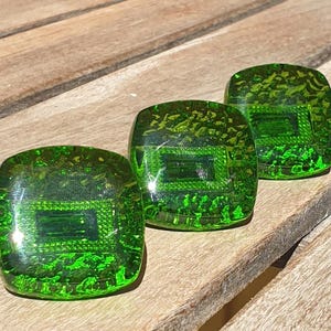 Könnte beinhalten: Drei quadratische, grüne Glasschrankknöpfe mit strukturierter Oberfläche und einem rechteckigen Design in der Mitte. Die Knöpfe sind diagonal auf einer Holzoberfläche angeordnet.