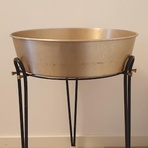 Può includere: Un vaso da fiori in metallo dorato con parte superiore rotonda e tre gambe in metallo nero. Il vaso è su un pavimento in legno chiaro.