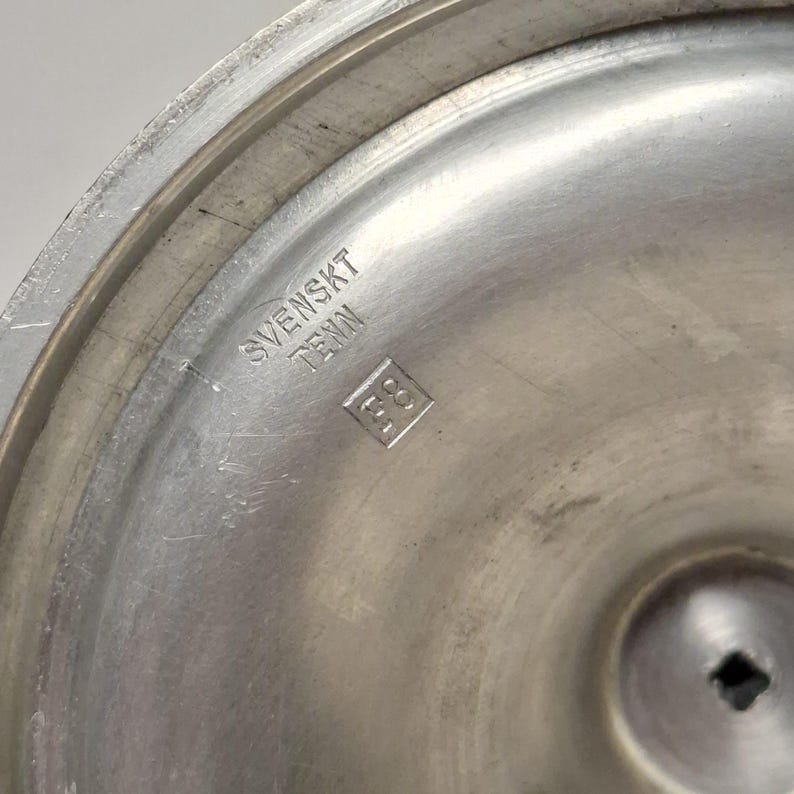 K&ouml;nnte beinhalten: Nahaufnahme eines silberfarbenen Metallobjekts mit dem Text "SVENSKT TENN" und einer quadratischen Markierung. Das Objekt hat eine kreisf&ouml;rmige Form mit leicht strukturierter Oberfl&auml;che und einem zentralen quadratischen Loch.