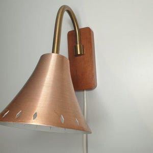 May include: A mid-century modern wall sconce with a copper-colored shade and a brass-colored curved arm. The shade has diamond-shaped cutouts. The sconce is mounted on a rectangular wooden base.