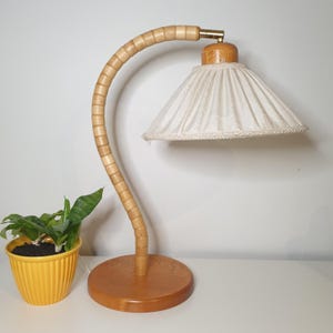 Może przedstawiać: Lampa biurkowa w stylu vintage z zakrzywionym, jasnym drewnianym trzonem i okrągłą podstawą. Lampa posiada kremowy abażur z ozdobnym wykończeniem. Mała żółta doniczka z zieloną rośliną stoi obok lampy.