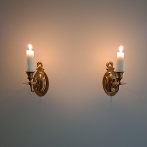May include: A pair of polished brass wall sconces, each featuring an oval backplate with a circular mounting hole at the top. Each sconce holds a single white pillar candle, with a lit flame casting a warm glow against the wall surface.