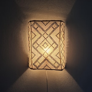 Könnte beinhalten: Eine rechteckige Wandleuchte mit einem beigen Häkelschirm und einem dunkelbraunen geometrischen Muster. Das Licht strahlt warm durch das komplexe, offene Netzgewebe und wirft deutliche Schatten an die Wand unter und neben der Leuchte.