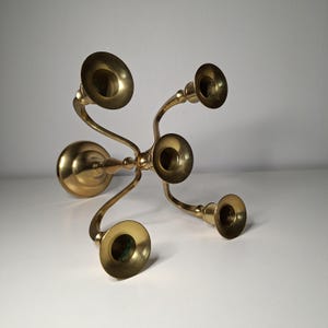 Può includere: Un candelabro in ottone lucido con cinque portacandele disposti a croce. Il supporto centrale è rialzato, con quattro bracci curvi che si estendono verso l'esterno per sostenere i restanti supporti, poggianti su una base circolare.