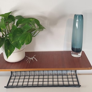 May include: A mid-century modern wall shelf with a dark wood top and a black wire grid base. A potted plant with green and yellow leaves, a blue glass vase, and a decorative metal object are displayed on the shelf.