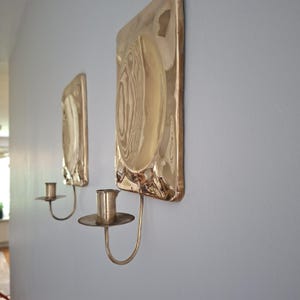 May include: Two wall-mounted candle holders made of polished brass. Each piece features a rectangular backplate with a central circular indentation and a smooth, reflective surface. A curved arm extends from the bottom to support a cylindrical candle cup.