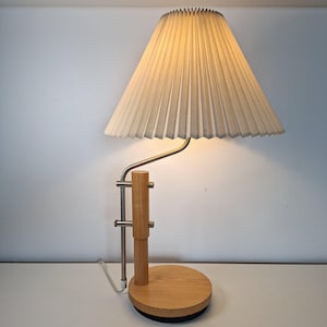 Może przedstawiać: Nowoczesna lampa stołowa z plisowanym abażurem. Lampa posiada jasną drewnianą podstawę i metalowe ramię, które podtrzymuje abażur. Klosz jest w jasnobeżowym kolorze, a lampa jest włączona, emitując ciepłą poświatę.