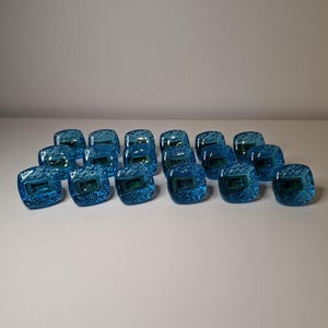 May include: A collection of square, blue glass cabinet knobs with a textured surface and a rectangular green design in the center. The knobs are arranged on a white surface.