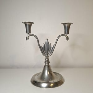 K&ouml;nnte beinhalten: Ein silberfarbener, zweiarmiger Kerzenleuchter mit einem dekorativen Blattdesign in der Mitte. Der Kerzenleuchter hat zwei Kerzenhalter und einen runden Sockel. Das Metall hat eine matte Oberfl&auml;che und wirkt dadurch vintage.