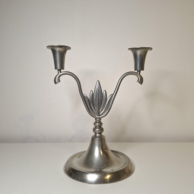 K&ouml;nnte beinhalten: Ein silberfarbener, zweiarmiger Kerzenleuchter mit einem dekorativen Blattdesign in der Mitte. Der Kerzenleuchter hat zwei Kerzenhalter und einen runden Sockel. Das Metall hat eine matte Oberfl&auml;che und wirkt dadurch vintage.