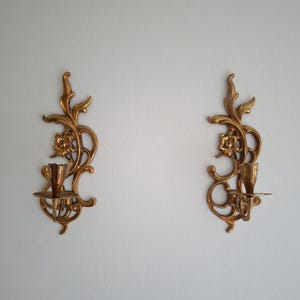 May include: A pair of matching gold-toned metal wall sconces featuring an intricate, ornate design with swirling leaf patterns and a central flower motif. Each sconce includes a small, circular candle holder platform extending from the base.