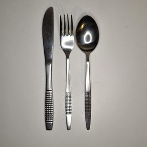 Op de afbeelding: Een roestvrijstalen bestekset bestaande uit een mes, vork en lepel. De handvatten hebben een dambordpatroon en de items zijn gerangschikt op een witte ondergrond. Het mes staat links, de vork in het midden en de lepel rechts.