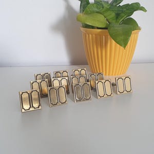 Puede incluir: Un conjunto de gemelos rectangulares dorados con borde negro y dos rectángulos verticales. Están dispuestos sobre una superficie blanca. Un jarrón amarillo acanalado con una planta verde se encuentra en el fondo.