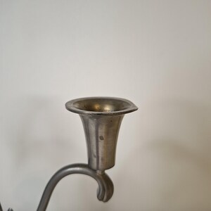 K&ouml;nnte beinhalten: Nahaufnahme eines Kerzenhalters aus Zinn. Der Kerzenhalter hat eine ausgestellte, becherf&ouml;rmige Oberseite und einen gebogenen Arm. Das Metall hat ein stumpfes, gealtertes Aussehen, was darauf hindeutet, dass es antik oder vintage ist.