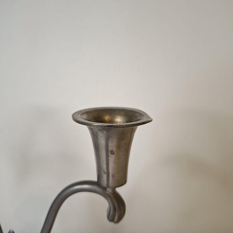 K&ouml;nnte beinhalten: Nahaufnahme eines Kerzenhalters aus Zinn. Der Kerzenhalter hat eine ausgestellte, becherf&ouml;rmige Oberseite und einen gebogenen Arm. Das Metall hat ein stumpfes, gealtertes Aussehen, was darauf hindeutet, dass es antik oder vintage ist.