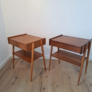 Könnte beinhalten: Zwei Nachttische aus Holz im Mid-Century-Modern-Stil. Jeder Tisch hat eine rechteckige Oberseite, eine Schublade und ein unteres Regal. Die Tische werden von abgewinkelten, sich verjüngenden Beinen getragen. Das Holz hat eine warme, natürliche Oberfläche.