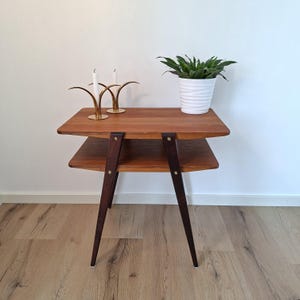 May include: A mid-century modern two-tiered side table with a warm wood finish. The table has dark brown legs and supports. Two gold candle holders with white candles and a white potted plant sit on the top shelf.