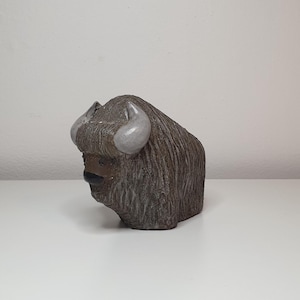 May include: A ceramic sculpture of a brown and gray musk ox with white horns. The sculpture is in a seated position with its head lowered.