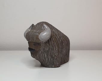 May include: A ceramic sculpture of a brown and gray musk ox with white horns. The sculpture is in a seated position with its head lowered.
