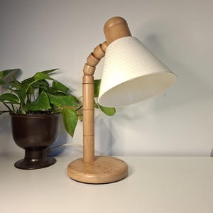 Może przedstawiać: Drewniana lampa biurkowa z białym abażurem. Lampa ma okrągłą podstawę i segmentowane ramię do regulacji. Klosz ma kształt stożka i jest wykonany z białej tkaniny o fakturze. Roślina doniczkowa znajduje się w tle.