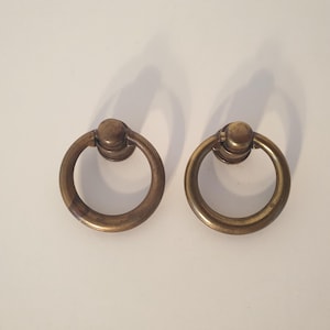 May include: A pair of antique brass ring pulls. Each pull consists of a circular ring connected to a decorative base. The brass has a warm, aged patina, indicating a vintage aesthetic. These pulls are designed for easy gripping and opening of drawers or cabinets.