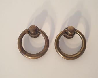 May include: Two antique brass ring pulls for furniture. Each pull features a circular ring attached to a decorative base. The brass has a warm, aged patina, suggesting vintage or retro styling. The pulls are designed for easy gripping and opening of drawers or cabinets.