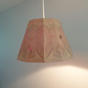 Può includere: Paralume geometrico beige con motivo a venature del legno. Il paralume è sospeso da un cavo bianco e presenta una serie di riflessi rosso-rosati. Il design presenta una serie di cerchi concentrici.