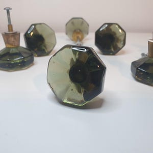 Könnte beinhalten: Eine Sammlung von olivgrünen Glasschrankknöpfen mit achteckigem Facettendesign. Jeder Knopf hat eine Metallschraube und einen Korkfuß. Die Knöpfe sind auf einer weißen Oberfläche angeordnet.