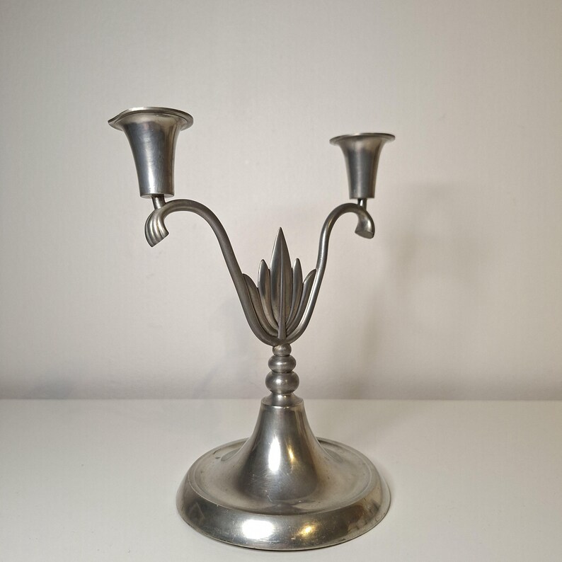 K&ouml;nnte beinhalten: Ein silberfarbener, zweiarmiger Kerzenhalter mit dekorativem Blattdesign. Der Sockel ist rund, und die Arme kr&uuml;mmen sich nach oben, um die Kerzen zu halten. Der Kerzenhalter ist aus Metall und hat eine Vintage-&Auml;sthetik.