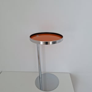 May include: A modern side table with a round, orange tabletop and a chrome-finished metal frame. The table has a circular base and a single, straight leg supporting the tabletop. The tabletop is approximately 16 inches in diameter.