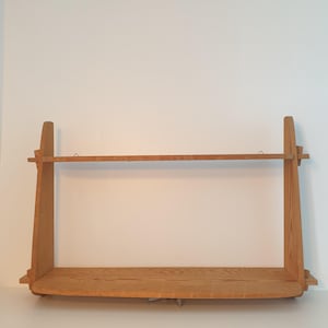 May include: A light brown wooden shelf with a horizontal bar and a lower shelf. The shelf has a simple, minimalist design with a natural wood grain finish. The shelf is designed to be mounted on a wall.
