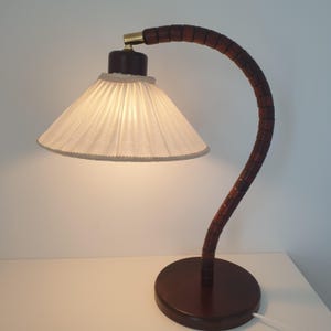 Może przedstawiać: Lampka biurkowa w stylu vintage z ciemnobrązową drewnianą podstawą i zakrzywionym, segmentowym drewnianym ramieniem. Lampa posiada biały, plisowany abażur z ozdobnym wykończeniem. Metalowe akcenty lampy są w kolorze złotym.