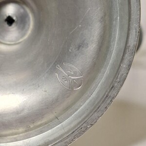 K&ouml;nnte beinhalten: Nahaufnahme eines silberfarbenen Metallobjekts mit kreisf&ouml;rmiger Basis und einem leicht erh&ouml;hten Mittelbereich. Das Objekt hat eine matte Oberfl&auml;che und ein kleines, eingraviertes Emblem in der N&auml;he der Mitte. Das Emblem scheint ein stilisiertes Design zu sein.