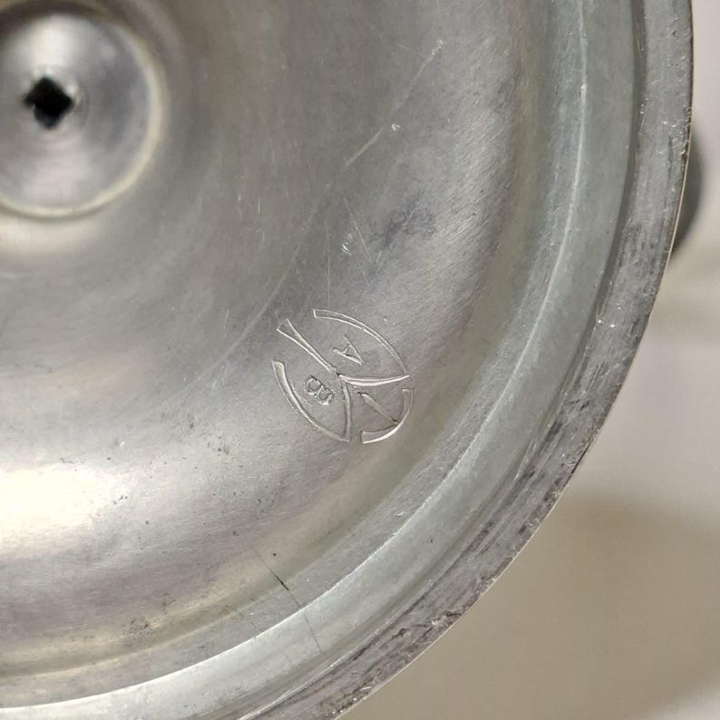K&ouml;nnte beinhalten: Nahaufnahme eines silberfarbenen Metallobjekts mit kreisf&ouml;rmiger Basis und einem leicht erh&ouml;hten Mittelbereich. Das Objekt hat eine matte Oberfl&auml;che und ein kleines, eingraviertes Emblem in der N&auml;he der Mitte. Das Emblem scheint ein stilisiertes Design zu sein.
