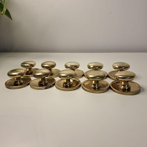 May include: A set of ten vintage brass cabinet knobs. Each knob has a rounded top and a circular base. The knobs are arranged in two rows on a white surface. A few green leaves are visible in the background.