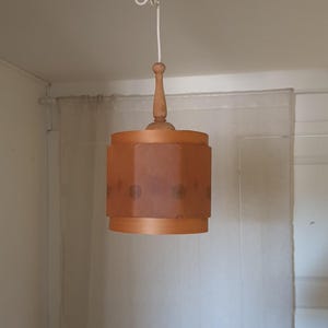 May include: A vintage wooden pendant light fixture. The light has a cylindrical shade with a warm, orange hue and a decorative pattern. The fixture is suspended from the ceiling by a white cord and a wooden rod.