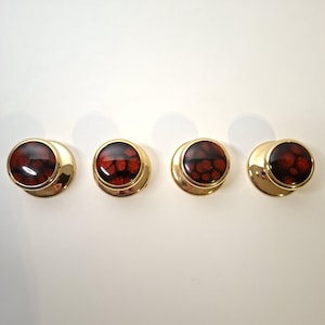 Puede incluir: Cuatro gemelos dorados con un diseño circular. Cada gemelo presenta un centro rojo oscuro moteado, encerrado en un marco de metal dorado. Los gemelos están dispuestos horizontalmente sobre un fondo blanco.