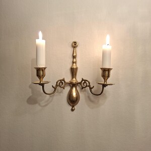 May include: A polished brass wall sconce featuring two arms with candle cups, each holding a white pillar candle. The central vertical support has a decorative finial at the top and a rounded base, with ornate scrollwork connecting the arms to the center.