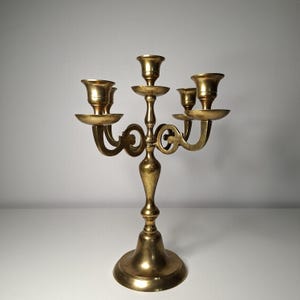 May include: A five-arm brass candelabra featuring a central candle holder flanked by four smaller holders on curved, scroll-shaped arms. The piece has a polished metallic finish, a tiered pedestal base, and a slender, tapered central column.