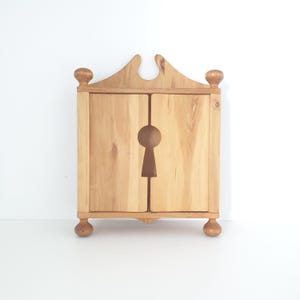 Könnte beinhalten: Kleiner, hellfarbiger Holzschrank mit Schlüsselloch-Design. Der Schrank hat zwei Türen und ist mit dekorativen Zierknöpfen oben und unten verziert. Die Holzmaserung ist sichtbar, und der Schrank steht vor einem weißen Hintergrund.