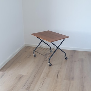 Puede incluir: Una mesa auxiliar de estilo moderno de mediados de siglo con una tapa rectangular de madera marrón. La mesa tiene una estructura de metal negro con un estante inferior y cuatro ruedas. La mesa mide aproximadamente 61 cm de largo.