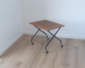 May include: A mid-century modern style side table with a rectangular, brown wooden top. The table has a black metal frame with a lower shelf and four rolling wheels. The table is approximately 24 inches long.