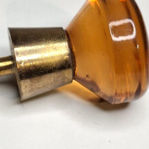 Op de afbeelding: Close-up van een vintage amberkleurige glazen kastknop met een messing voet. De knop heeft een afgeronde vorm en een warme, doorschijnende kleur. De messing voet vertoont tekenen van ouderdom.