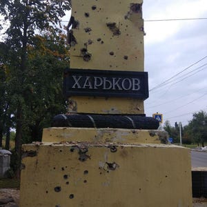 May include: A yellow concrete obelisk with the word "ХАРЬКОВ" in black lettering on a black plaque. The obelisk is covered in bullet holes.