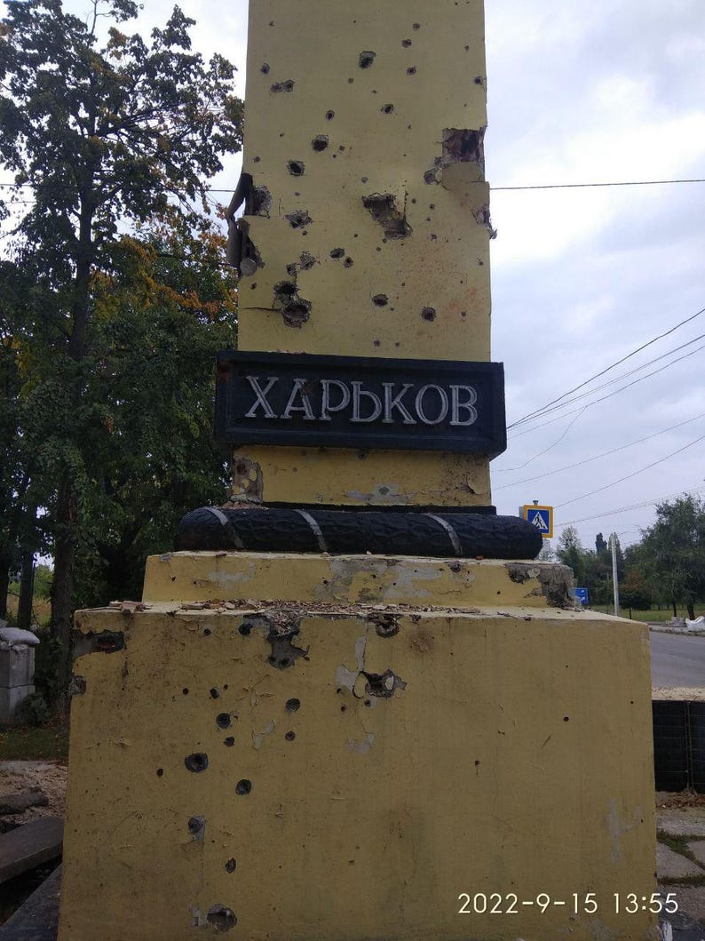 May include: A yellow concrete obelisk with the word "ХАРЬКОВ" in black lettering on a black plaque. The obelisk is covered in bullet holes.