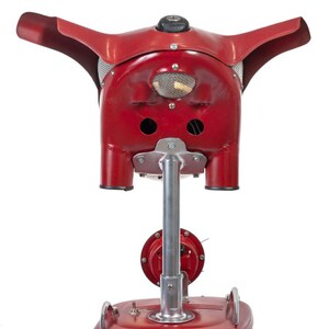 May include: A red vintage electric heater with a unique design resembling a pig's head. The heater has a metal base and a long metal pole that supports the head. The head has two large holes on the sides and a small mesh grill on the front.